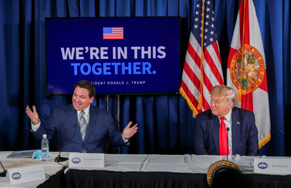 Ron DeSantis and Donald Trump sit at a table before microphones in front of flags and sign reading: We're in this together, President Donald J. Trump