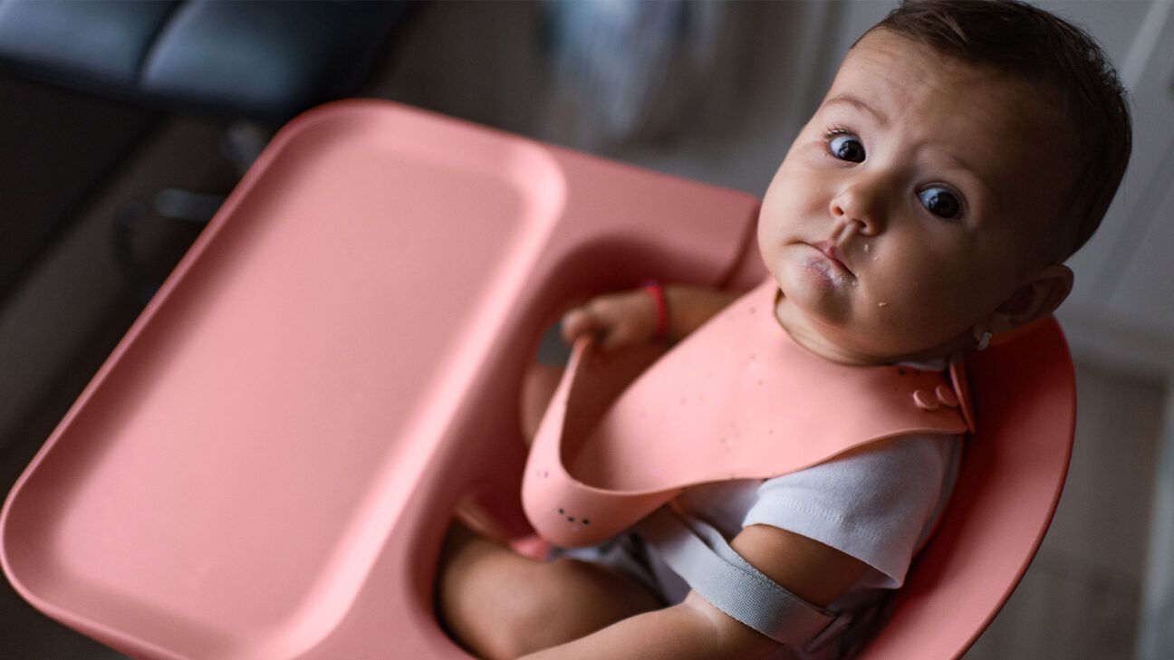 A baby in a pink high chair looks at the camera.