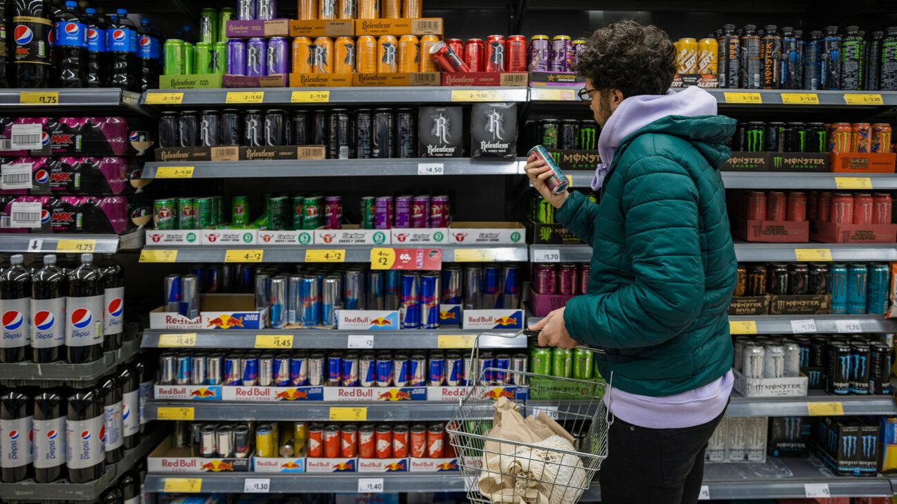 Man in green jacket picks up canned beverage at store.